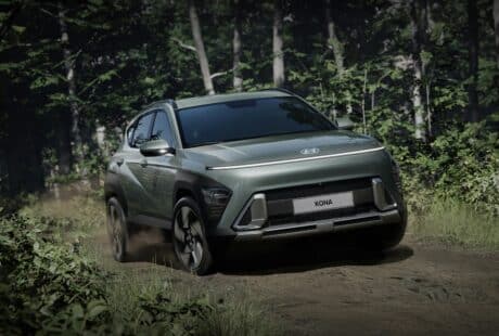 A metallic green Hyundai Kona SUV drives on a dirt track through a lush, green forest. The car kicks up dust and sunlight filters through dense trees in the background, highlighting the Kona’s sleek, modern design and prominent front grille.
