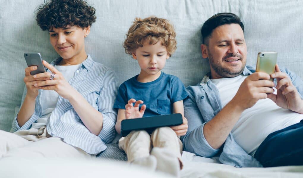 Three people sit on a sofa: an adult on the left using a mobile, a child in the middle using a tablet, and an adult on the right also using a mobile. All are focused on their devices and appear relaxed, wearing casual clothes.