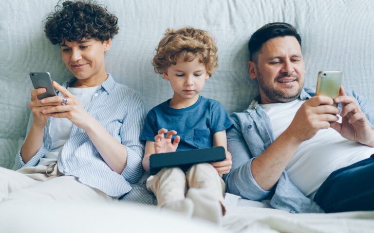 Three people sit on a sofa: an adult on the left using a mobile, a child in the middle using a tablet, and an adult on the right also using a mobile. All are focused on their devices and appear relaxed, wearing casual clothes.