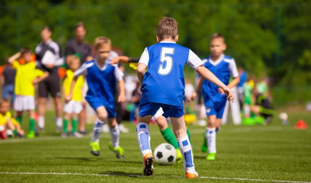 A youth football match in progress. A boy wearing a blue shirt with number 5 is running towards a black-and-white football, surrounded by teammates and opponents, all wearing sports kits. Spectators and greenery are visible in the blurred background.