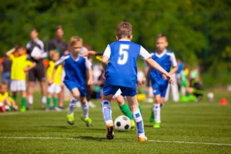 A youth football match in progress. A boy wearing a blue shirt with number 5 is running towards a black-and-white football, surrounded by teammates and opponents, all wearing sports kits. Spectators and greenery are visible in the blurred background.