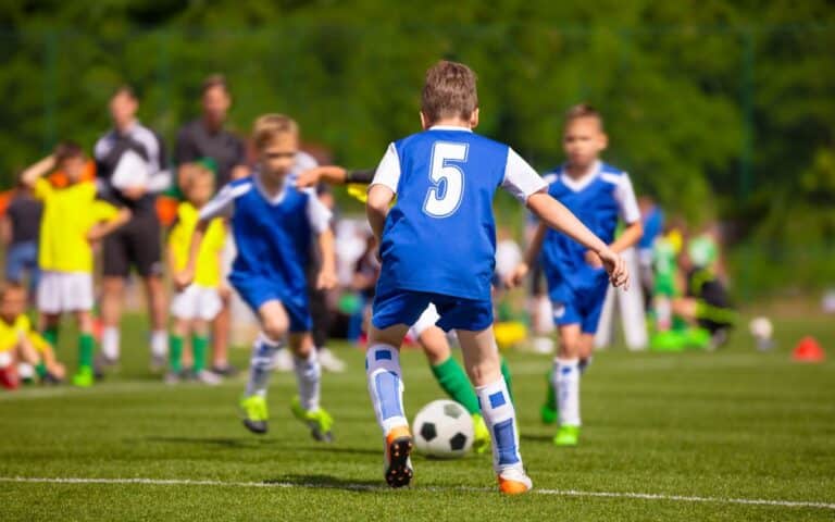 A youth football match in progress. A boy wearing a blue shirt with number 5 is running towards a black-and-white football, surrounded by teammates and opponents, all wearing sports kits. Spectators and greenery are visible in the blurred background.