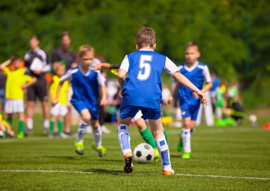A youth football match in progress. A boy wearing a blue shirt with number 5 is running towards a black-and-white football, surrounded by teammates and opponents, all wearing sports kits. Spectators and greenery are visible in the blurred background.