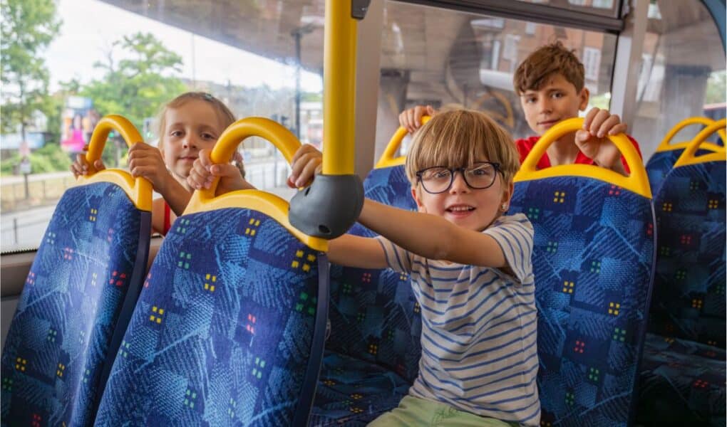 Three children sit on blue patterned seats inside a bus, holding yellow handles. The boy in front wears glasses and a striped shirt, smiling at the camera, whilst the two children behind him look forward. Trees and buildings are visible through the bus windows.