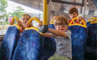 Three children sit on blue patterned seats inside a bus, holding yellow handles. The boy in front wears glasses and a striped shirt, smiling at the camera, whilst the two children behind him look forward. Trees and buildings are visible through the bus windows.