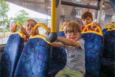 Three children sit on blue patterned seats inside a bus, holding yellow handles. The boy in front wears glasses and a striped shirt, smiling at the camera, whilst the two children behind him look forward. Trees and buildings are visible through the bus windows.