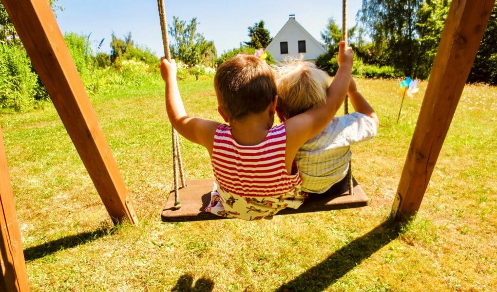 Two young children sit side by side on a wooden swing, facing a sunlit grassy garden. The child on the left wears a red striped top, and the other wears a pale shirt. Trees, flowers, and a small white house are visible in the bright background.