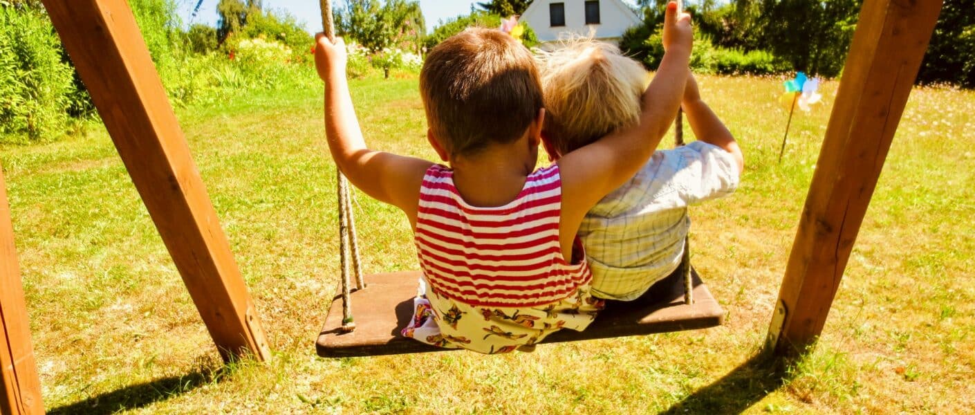 Two young children sit side by side on a wooden swing, facing a sunlit grassy garden. The child on the left wears a red striped top, and the other wears a pale shirt. Trees, flowers, and a small white house are visible in the bright background.