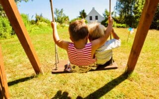 Two young children sit side by side on a wooden swing, facing a sunlit grassy garden. The child on the left wears a red striped top, and the other wears a pale shirt. Trees, flowers, and a small white house are visible in the bright background.