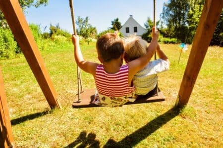 Two young children sit side by side on a wooden swing, facing a sunlit grassy garden. The child on the left wears a red striped top, and the other wears a pale shirt. Trees, flowers, and a small white house are visible in the bright background.