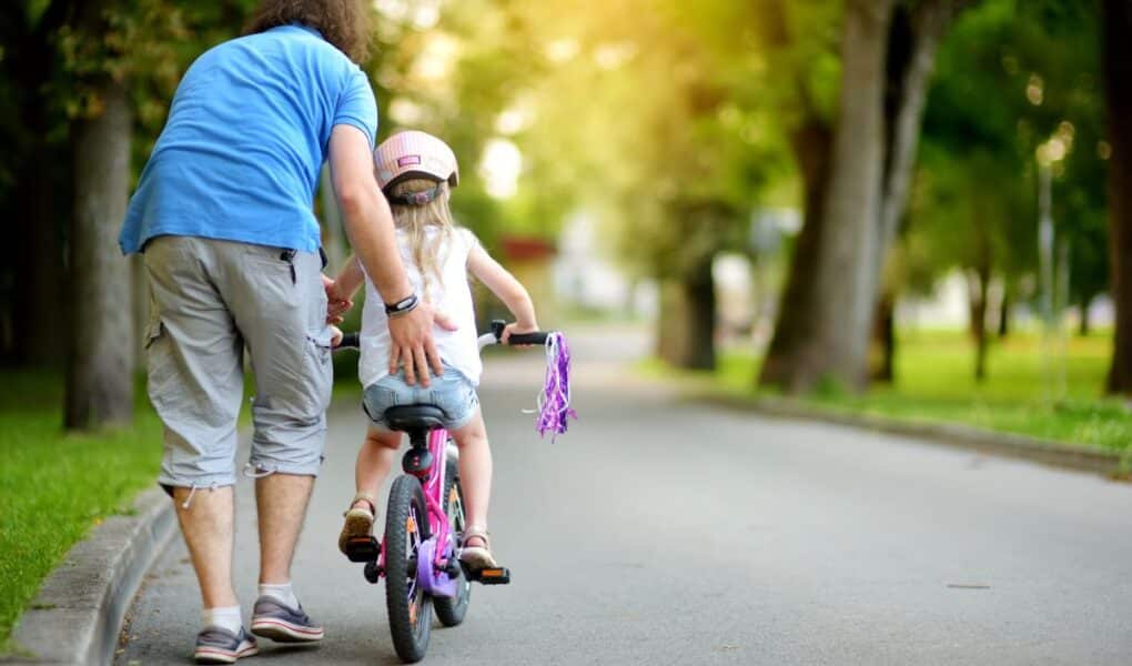 An adult helps a young child ride a bike with stabilisers on a paved path in a park. The adult wears a blue shirt and grey shorts, while the child wears a helmet. Trees and greenery line the path, with sunlight filtering through the leaves.
