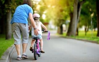 An adult helps a young child ride a bike with stabilisers on a paved path in a park. The adult wears a blue shirt and grey shorts, while the child wears a helmet. Trees and greenery line the path, with sunlight filtering through the leaves.