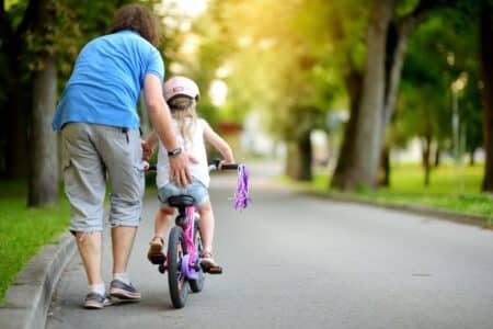 An adult helps a young child ride a bike with stabilisers on a paved path in a park. The adult wears a blue shirt and grey shorts, while the child wears a helmet. Trees and greenery line the path, with sunlight filtering through the leaves.
