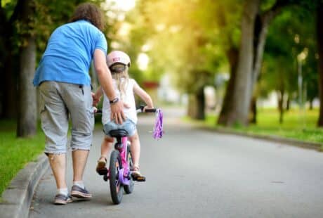 An adult helps a young child ride a bike with stabilisers on a paved path in a park. The adult wears a blue shirt and grey shorts, while the child wears a helmet. Trees and greenery line the path, with sunlight filtering through the leaves.
