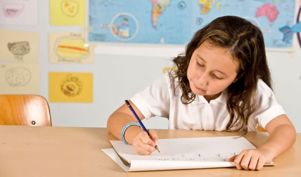 A young girl with wavy brown hair, wearing a white polo shirt, sits at a desk and writes in a notebook. Behind her are colourful drawings, a world map, and a blue wall. She looks focused on her work.