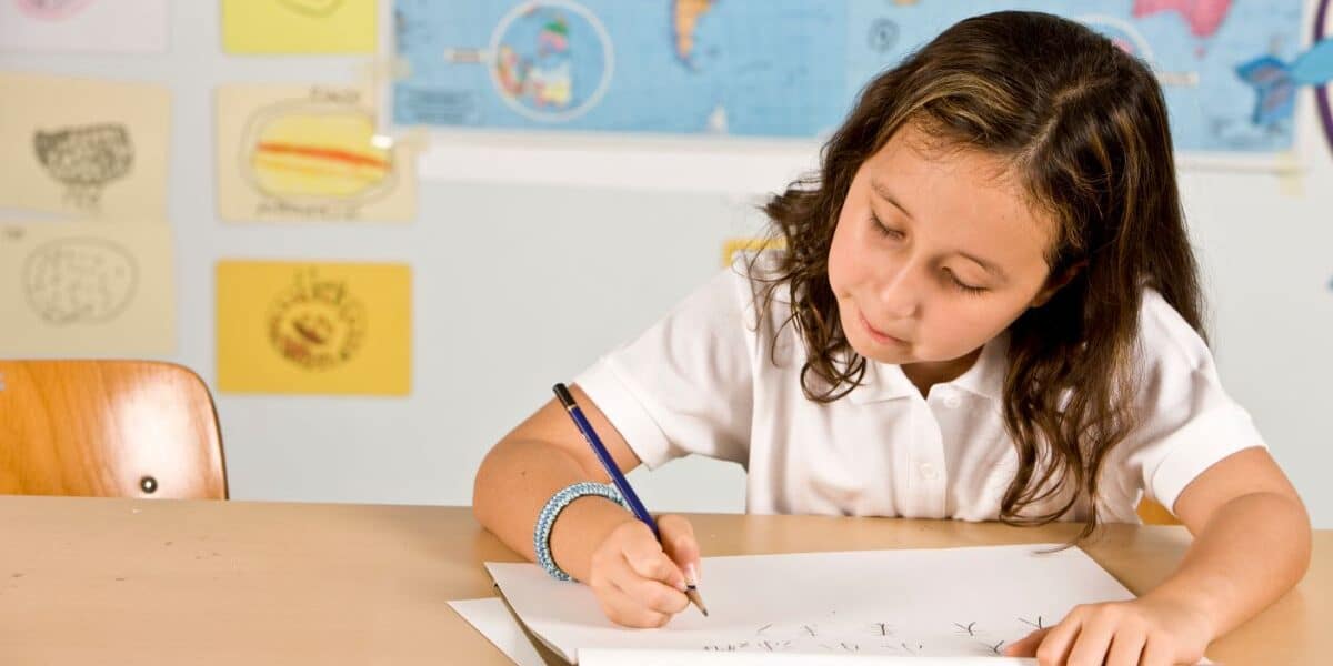 A young girl with wavy brown hair, wearing a white polo shirt, sits at a desk and writes in a notebook. Behind her are colourful drawings, a world map, and a blue wall. She looks focused on her work.