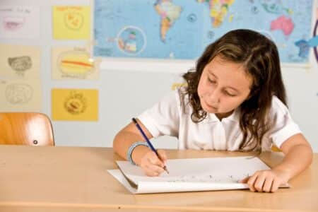 A young girl with wavy brown hair, wearing a white polo shirt, sits at a desk and writes in a notebook. Behind her are colourful drawings, a world map, and a blue wall. She looks focused on her work.
