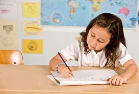 A young girl with wavy brown hair, wearing a white polo shirt, sits at a desk and writes in a notebook. Behind her are colourful drawings, a world map, and a blue wall. She looks focused on her work.