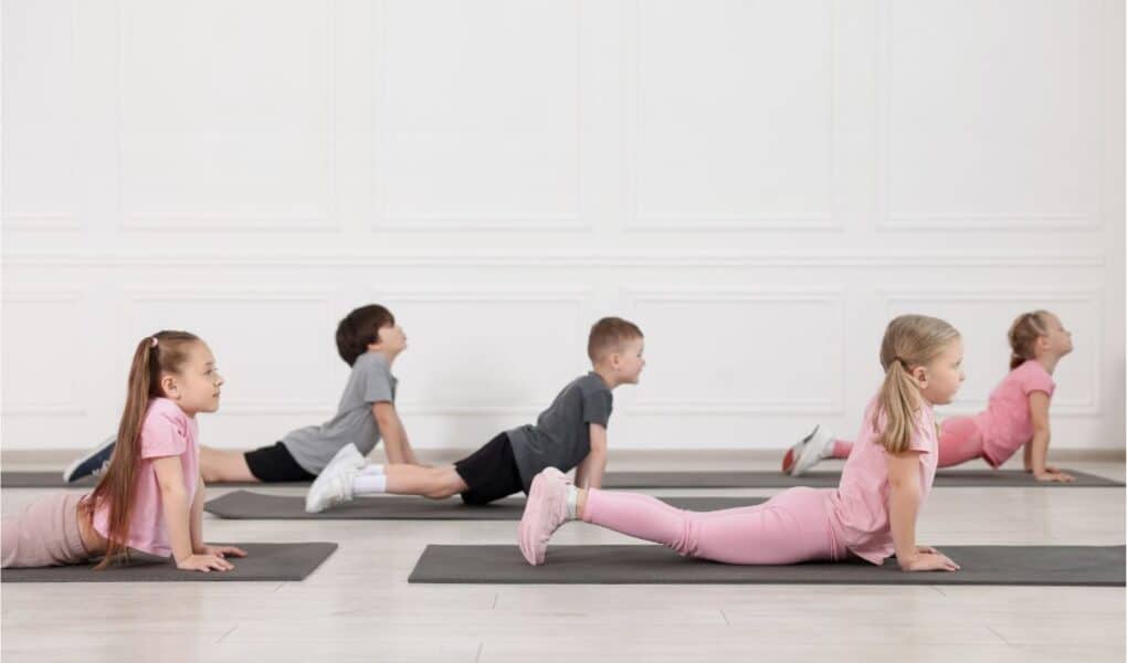 Five young children, three girls and two boys, are doing the upward-facing dog yoga pose on black mats in a bright room with white walls. All face the same direction, stretching their backs, with girls dressed in pink and boys in grey sportswear.