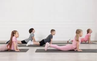 Five young children, three girls and two boys, are doing the upward-facing dog yoga pose on black mats in a bright room with white walls. All face the same direction, stretching their backs, with girls dressed in pink and boys in grey sportswear.