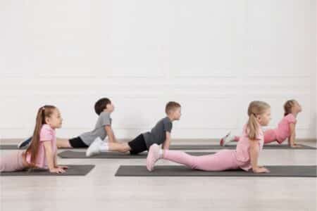 Five young children, three girls and two boys, are doing the upward-facing dog yoga pose on black mats in a bright room with white walls. All face the same direction, stretching their backs, with girls dressed in pink and boys in grey sportswear.