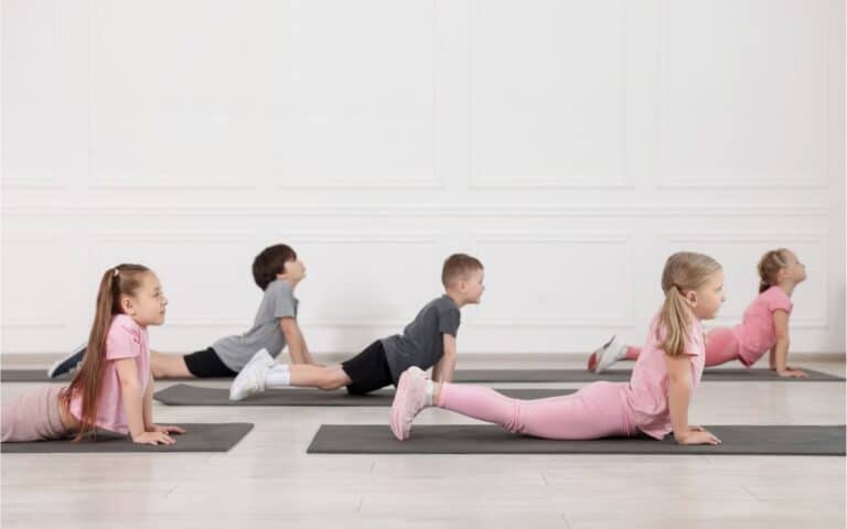 Five young children, three girls and two boys, are doing the upward-facing dog yoga pose on black mats in a bright room with white walls. All face the same direction, stretching their backs, with girls dressed in pink and boys in grey sportswear.
