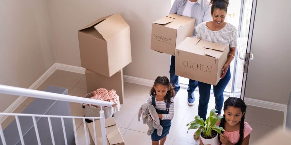 A family of four carries cardboard boxes and belongings into a new house. The parents, wearing casual clothes, carry boxes labelled “Bedroom” and “Kitchen,” while their two young daughters carry a soft toy and a potted plant as they walk up the hall stairs.
