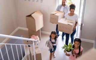 A family of four carries cardboard boxes and belongings into a new house. The parents, wearing casual clothes, carry boxes labelled “Bedroom” and “Kitchen,” while their two young daughters carry a soft toy and a potted plant as they walk up the hall stairs.