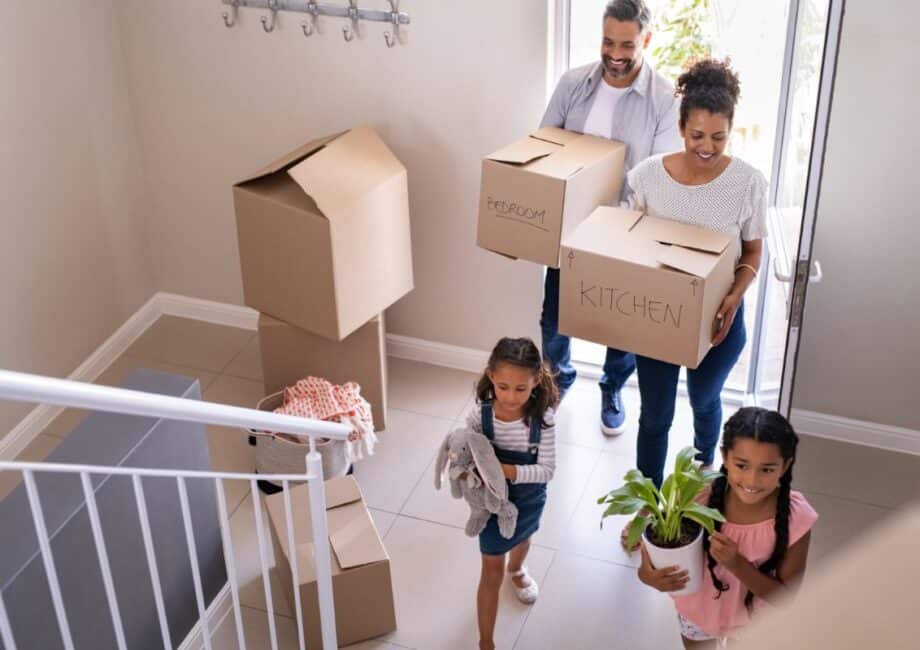 A family of four carries cardboard boxes and belongings into a new house. The parents, wearing casual clothes, carry boxes labelled “Bedroom” and “Kitchen,” while their two young daughters carry a soft toy and a potted plant as they walk up the hall stairs.