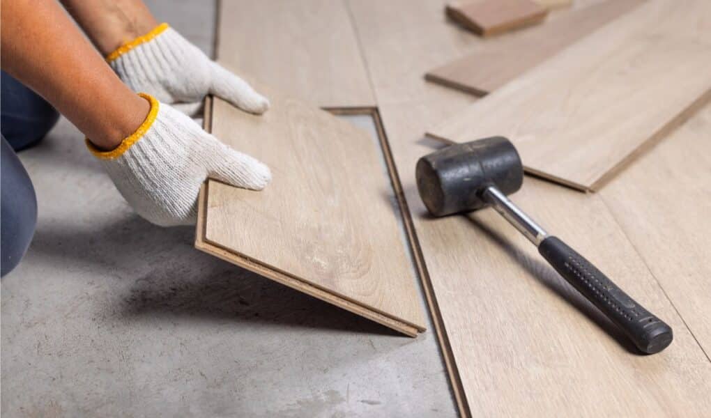 A person wearing white work gloves installs a light-coloured wood laminate floor plank. Nearby, a black rubber mallet lies on the partially finished floor, and other planks are scattered on the concrete subfloor.