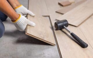 A person wearing white work gloves installs a light-coloured wood laminate floor plank. Nearby, a black rubber mallet lies on the partially finished floor, and other planks are scattered on the concrete subfloor.