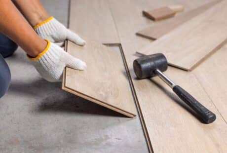 A person wearing white work gloves installs a light-coloured wood laminate floor plank. Nearby, a black rubber mallet lies on the partially finished floor, and other planks are scattered on the concrete subfloor.