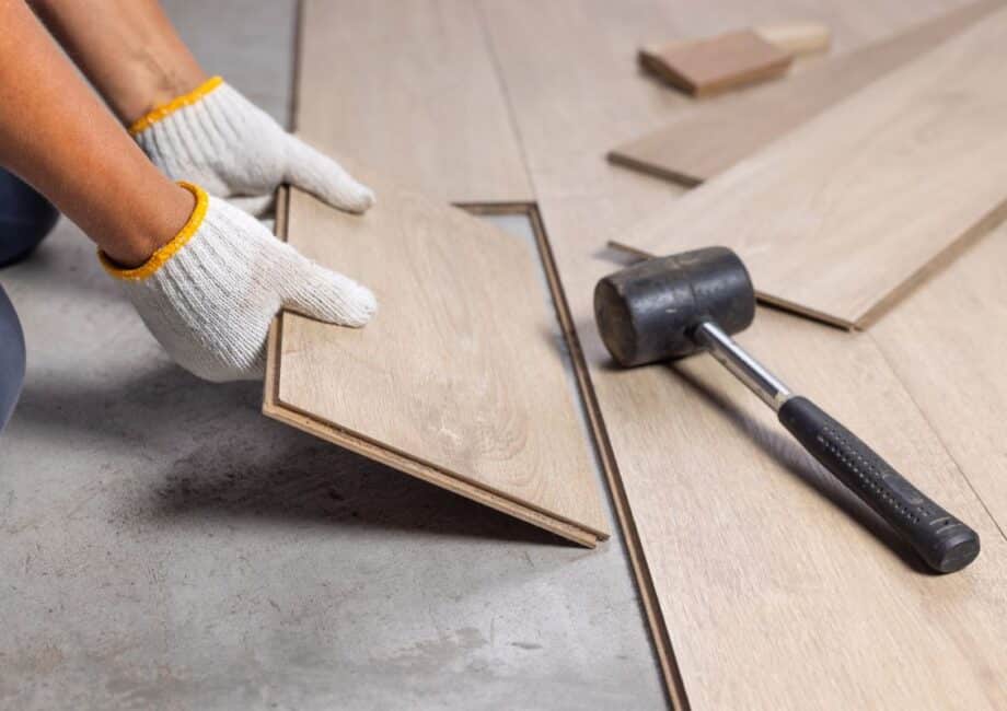 A person wearing white work gloves installs a light-coloured wood laminate floor plank. Nearby, a black rubber mallet lies on the partially finished floor, and other planks are scattered on the concrete subfloor.